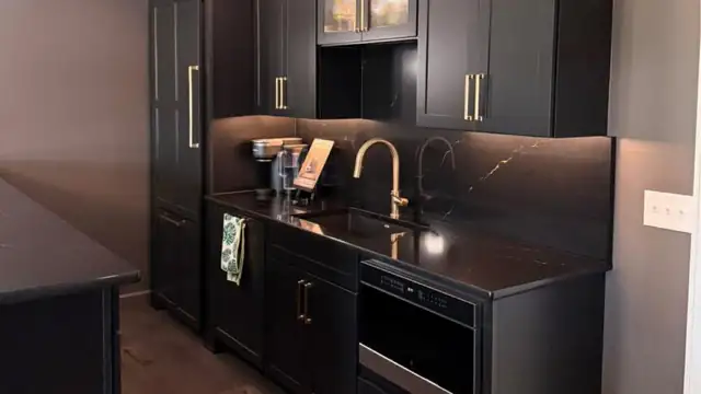 Black cabinets with dark marble-look countertop and backsplash, gold faucet and pulls, and under-cabinet lighting